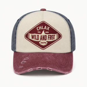 A distressed trucker-style baseball cap with a cream front panel, dark blue-gray side panels, and a washed burgundy brim. The front features a diamond-shaped embroidered patch with the text 'CHLAX', 'WILD AND FREE', and 'AMPM'.