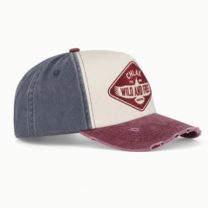A distressed two-tone baseball cap with a washed navy blue crown, an off-white front panel, and a faded burgundy brim. It features a diamond-shaped embroidered patch on the front with the text 'CHLAX WILD AND FREE'.