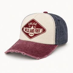 A distressed trucker-style baseball cap with a cream front panel, dark blue-gray side panels, and a faded burgundy brim. The front features a diamond-shaped embroidered patch with the text 'CHLAX TRK MK WILD AND FREE'.