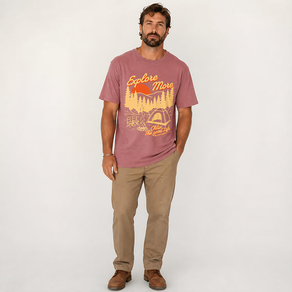 A man wearing a dusty rose graphic t-shirt with a yellow and orange outdoor design, paired with khaki pants and brown shoes, standing against a light gray studio background.