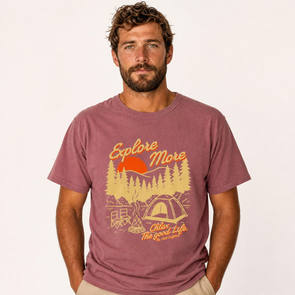 A man wearing a mauve-brown graphic t-shirt with a yellow and orange camping design that reads 'Explore More. CHLAX The good Life.