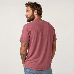 A man with curly brown hair and a beard, shown from the back wearing a dusty rose short-sleeve t-shirt and light blue jeans, against a plain off-white background.