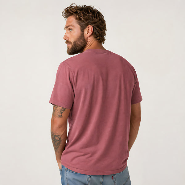 A man with curly brown hair and a beard, shown from the back wearing a dusty rose short-sleeve t-shirt and light blue jeans, against a plain off-white background.