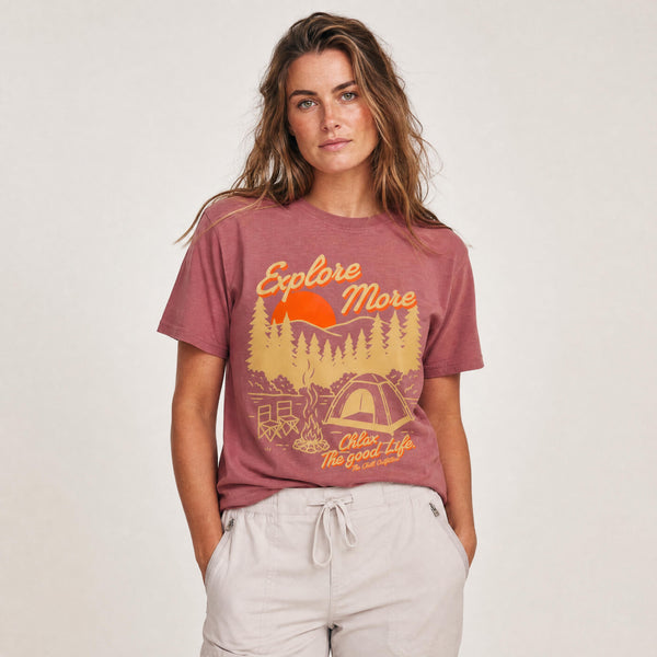A woman modeling a dusty rose short-sleeve graphic t-shirt with a yellow and orange camping design that reads 'Explore More' and 'CHLAX The Good Life'.