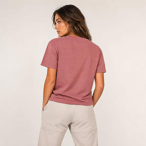 A woman shown from the back wearing a dusty rose short-sleeve t-shirt and light beige pants, photographed against a clean off-white studio background.