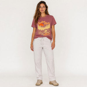 A woman modeling a dusty rose graphic t-shirt with a yellow and orange outdoor design, paired with light gray drawstring pants and beige sneakers, against a plain white studio background.