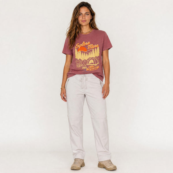 A woman modeling a dusty rose graphic t-shirt with a yellow and orange outdoor design, paired with light gray drawstring pants and beige sneakers, against a plain white studio background.