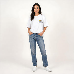 A woman modeling a white short-sleeve t-shirt with a small yellow and blue graphic on the left chest, paired with blue jeans and white sneakers, against a plain light background.