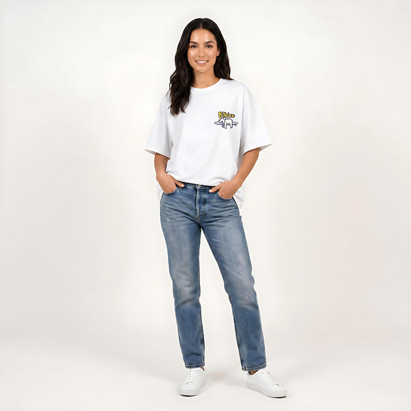A woman modeling a white short-sleeve t-shirt with a small yellow and blue graphic on the left chest, paired with blue jeans and white sneakers, against a plain light background.