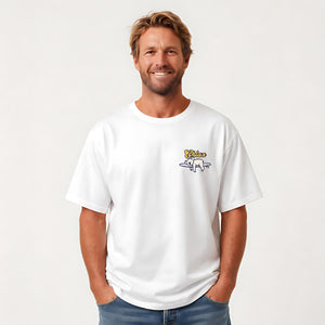 A smiling man wearing a white crewneck t-shirt with a small yellow and blue graphic on the left chest, paired with blue jeans, against a plain light background.