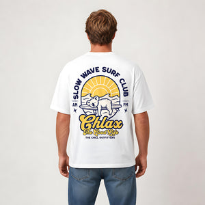 A man viewed from the back wearing a white t-shirt with a large graphic print on the back. The graphic features a yellow sun, a bear on a surfboard, and the text 'SLOW WAVE SURF CLUB' and 'Chlax The Good Life'.