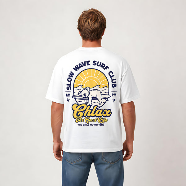 A man viewed from the back wearing a white t-shirt with a large graphic print on the back. The graphic features a yellow sun, a bear on a surfboard, and the text 'SLOW WAVE SURF CLUB' and 'Chlax The Good Life'.