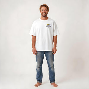 A man wearing a white crewneck t-shirt with a small graphic on the left chest and blue jeans, standing against a plain light gray studio background.