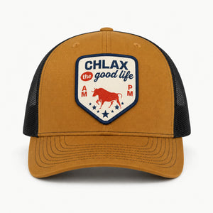 A tan and black trucker hat with a shield-shaped front patch that reads 'CHLAX the good life' with a red bull graphic and 'AM PM' text, shown on a light gray background.