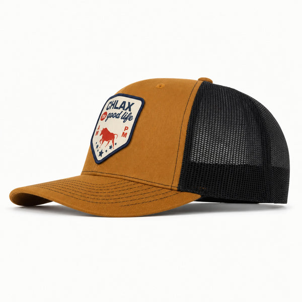 A trucker-style baseball cap with a camel brown front panel and brim, black mesh back, and a shield-shaped embroidered patch on the front that reads 'CHLAX good life TX' with a red bull graphic.