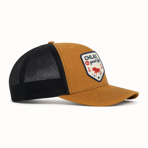 A trucker-style baseball cap with a mustard yellow front panel and brim, black mesh back, and a shield-shaped embroidered patch on the front that reads 'CHLAX is good life' with a red bull graphic.