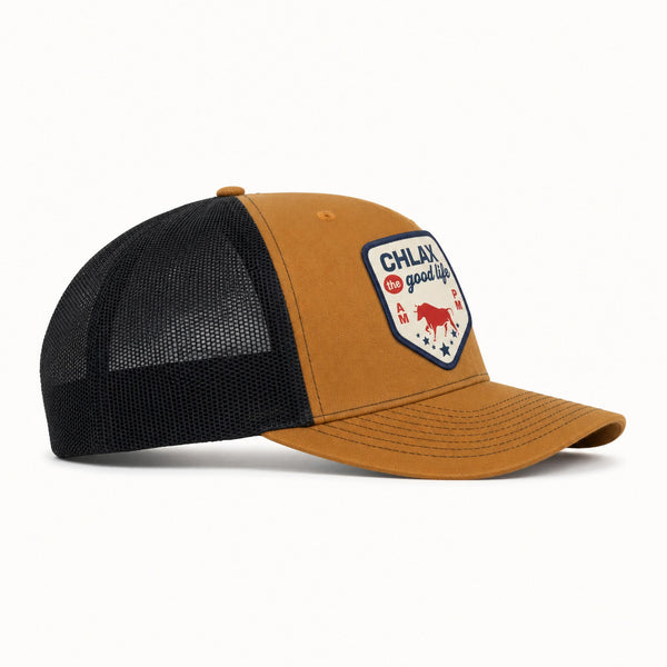A trucker-style baseball cap with a mustard yellow front panel and brim, black mesh back, and a shield-shaped embroidered patch on the front that reads 'CHLAX is good life' with a red bull graphic.