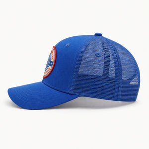 A royal blue trucker-style baseball cap with a curved brim, mesh back panels, and a circular embroidered patch on the side, shown against a plain white background.