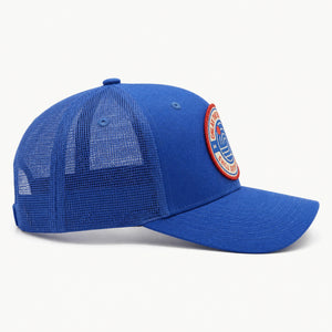 A royal blue trucker-style baseball cap with a mesh back and a circular embroidered patch on the side, photographed on a clean white background.