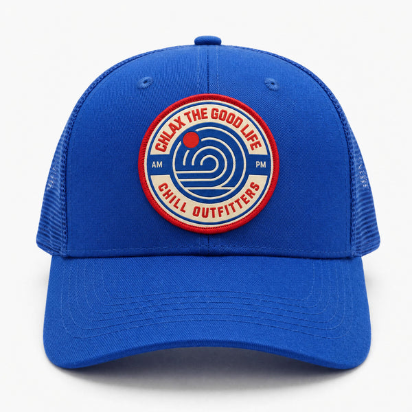 A royal blue trucker cap with a circular embroidered patch on the front. The patch features red and white text reading 'CHLAX THE GOOD LIFE CHILL OUTFITTERS' with a blue spiral logo in the center and 'AM PM' on either side.