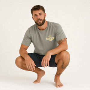 A man crouching in a studio setting, wearing a muted gray t-shirt with a yellow 'CHLAX WILD AND FREE' graphic on the left chest and dark shorts.