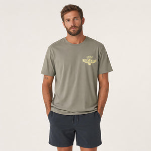 A man wearing a muted olive green t-shirt with a small yellow graphic on the left chest, paired with navy blue shorts, standing against a plain off-white studio background.