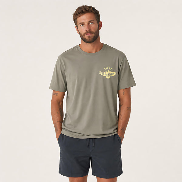 A man wearing a muted olive green t-shirt with a small yellow graphic on the left chest, paired with navy blue shorts, standing against a plain off-white studio background.