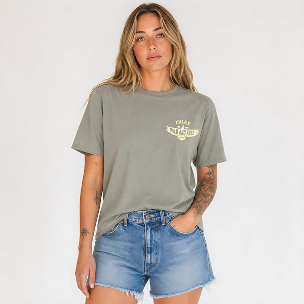 A woman modeling a muted sage green graphic t-shirt with a yellow 'CHLAX WILD AND FREE' print on the left chest, paired with light-wash denim shorts against a plain white background.
