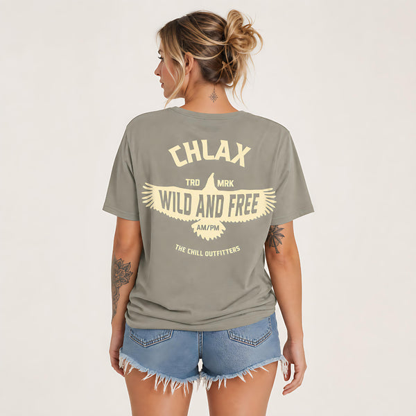 A woman shown from the back wearing an oversized olive green t-shirt with a cream graphic print that reads 'CHLAX' and 'WILD AND FREE'. She is also wearing denim cutoff shorts against a plain light background.
