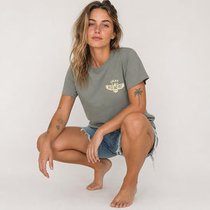 A woman crouching in a studio setting, wearing a muted olive green t-shirt with a yellow graphic print on the left chest and denim shorts.