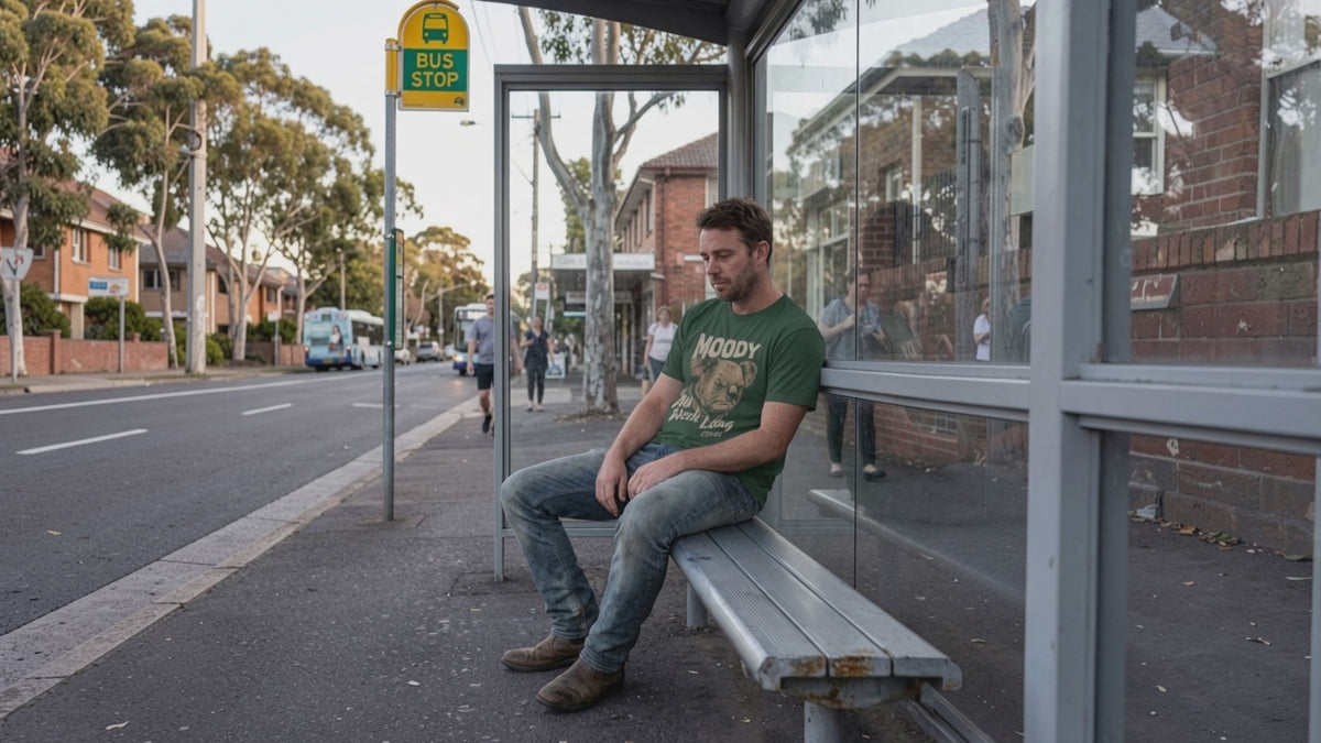 A man sitting at the bus stop watching the world go by wearing a green t-shirt with a moody koala on it.