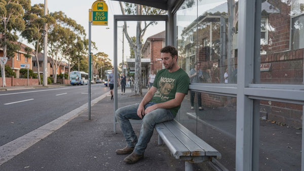 A man sitting at the bus stop watching the world go by wearing a green t-shirt with a moody koala on it.