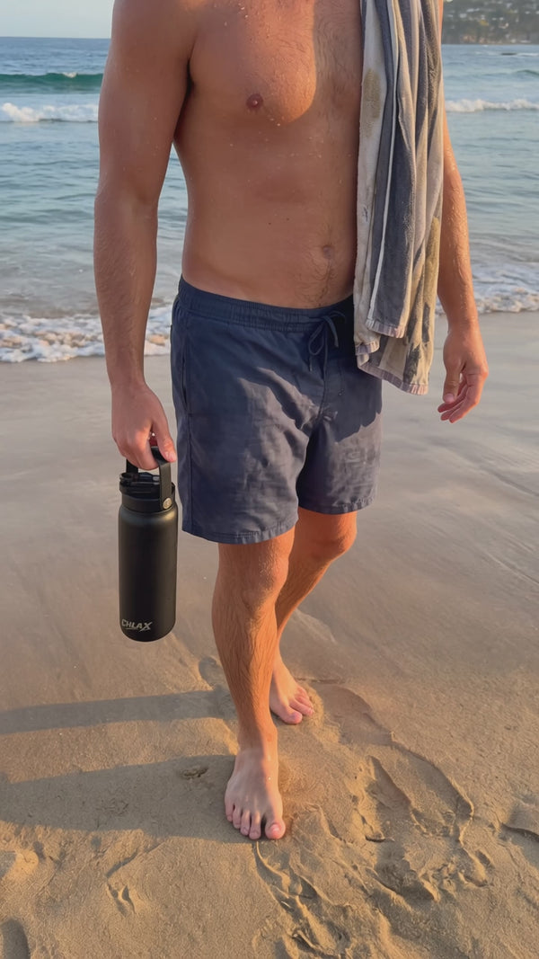 A man in blue boardshorts at the beach with a towel over the shoulder carrying a black CHLAX insulated drink bottle video.